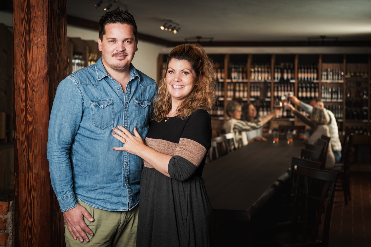 Michiel Jalink und Stephanie im Verkostungsraum der Brennerei Hödl Hof, im Hintergrund Regale mit Flaschen und Gäste beim Anstoßen