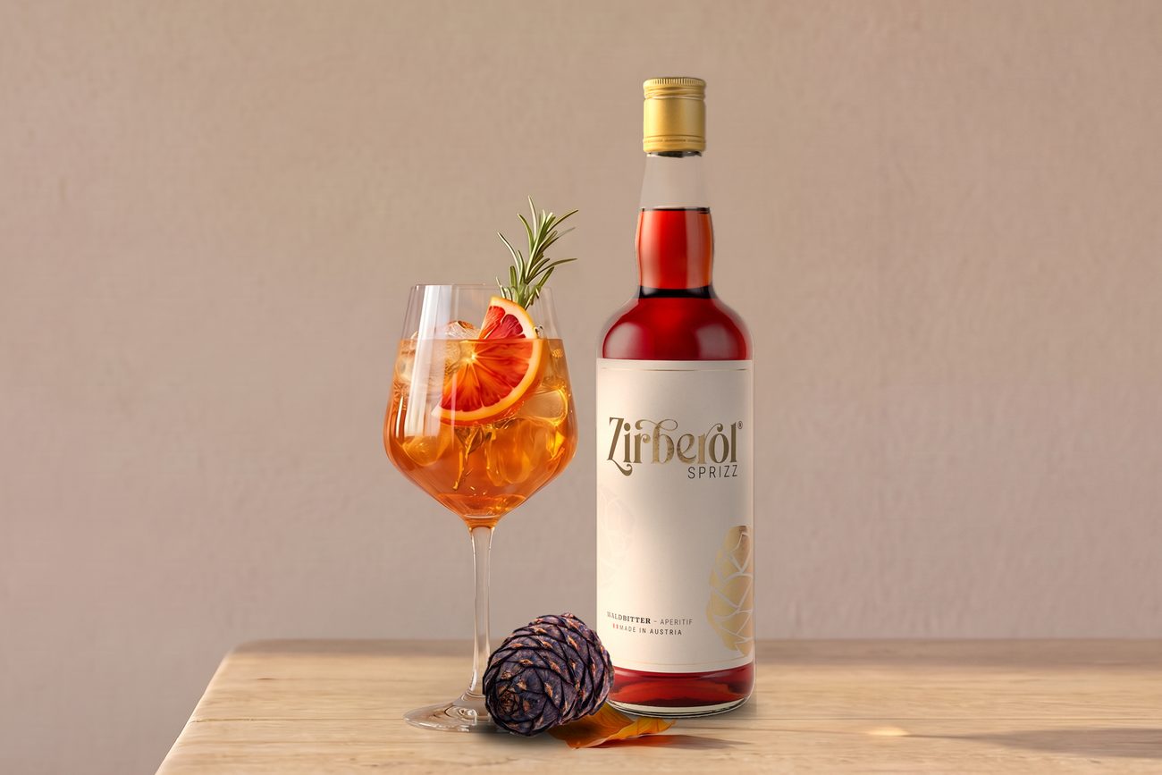 Zirberol Sprizz im Aperitifglas mit Blutorange und Rosmarin neben der Flasche und einem Zirbenzapfen